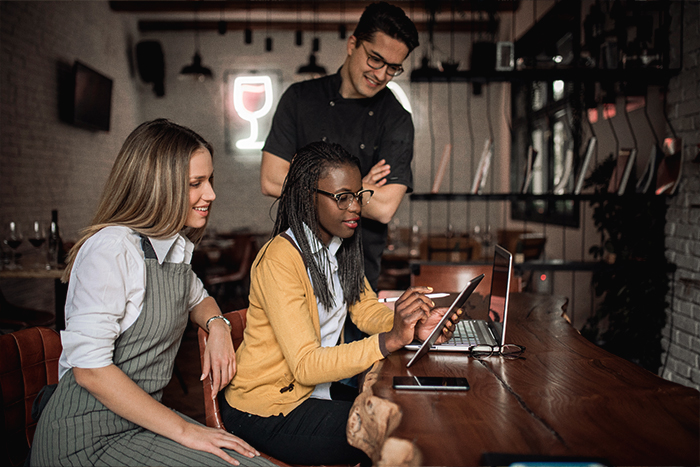 Three restaurant employees gathered around a tablet and a laptop in a restaurant.
