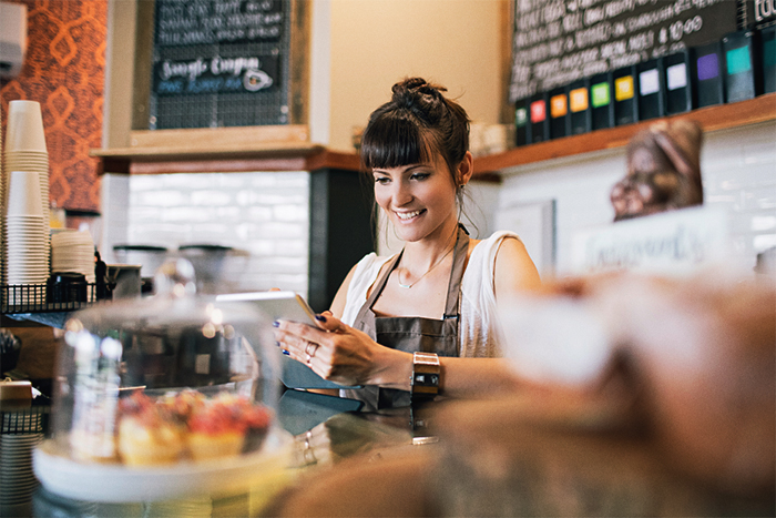 A woman standing behind a counter at a café smiling looking at a tablet.
