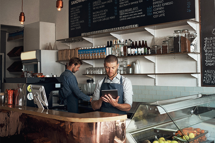 Two men behind a counter at a café, one is looking tablet and one is making coffee.