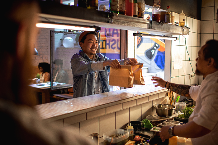 A man in a restaurant picking up two paper bags handed to him by a chef