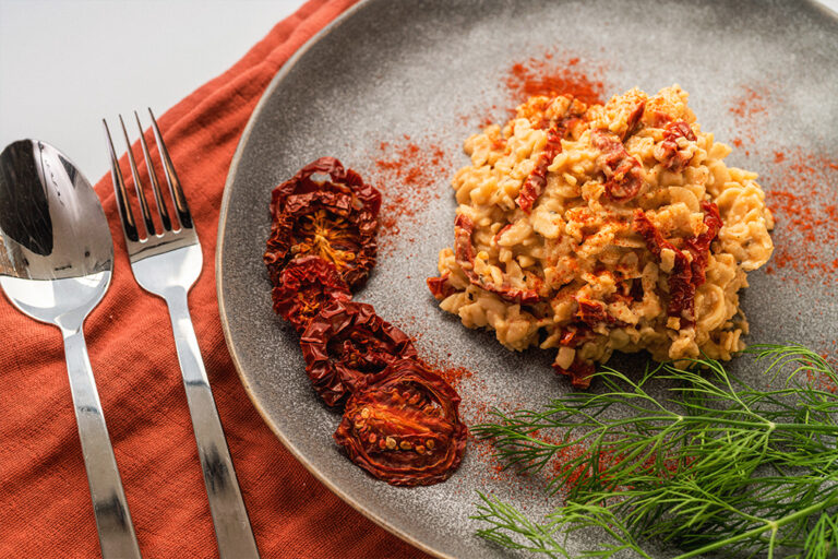 Pasta on a plate with roasted tomatoes and garnish beside a spoon and fork on a red placemat.