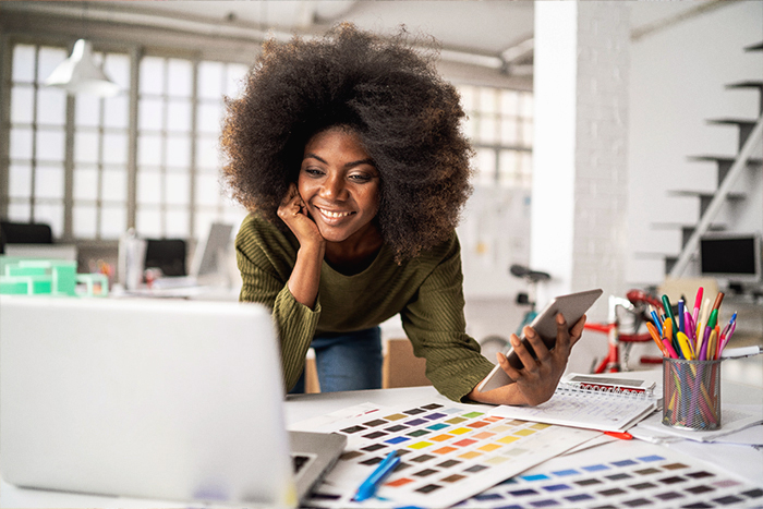 Woman smiling at a laptop screen holding a phone with colour palettes on a table in front of her.