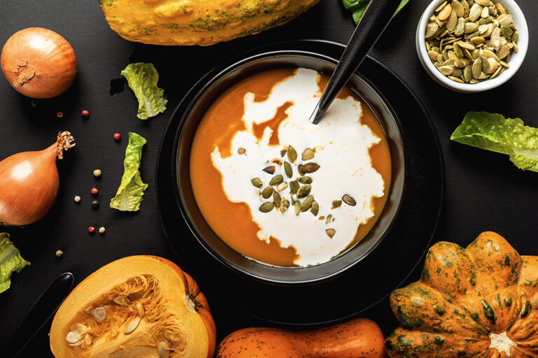 A black bowl of creamy pumpkin soup surrounded by a mix of colourful vegetables.